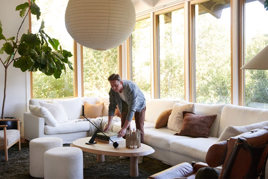 A man arranges books on a coffee table next to a white couch in a living room that has floor-to-ceiling picture windows with white oak frames.