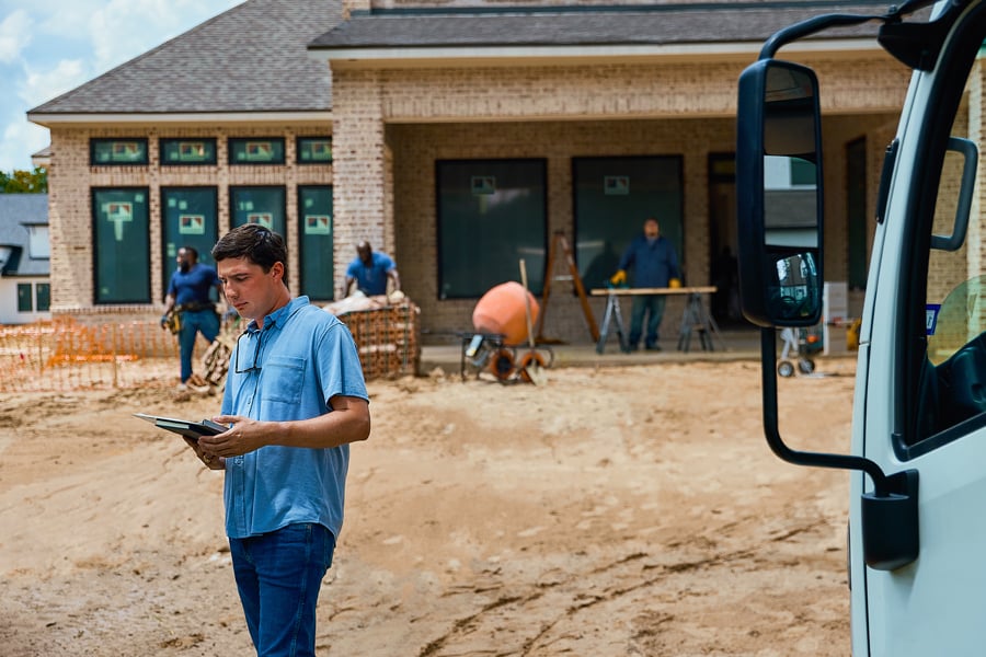 A man with a tablet stands on a construction site where windows are being installed in a new construction home.