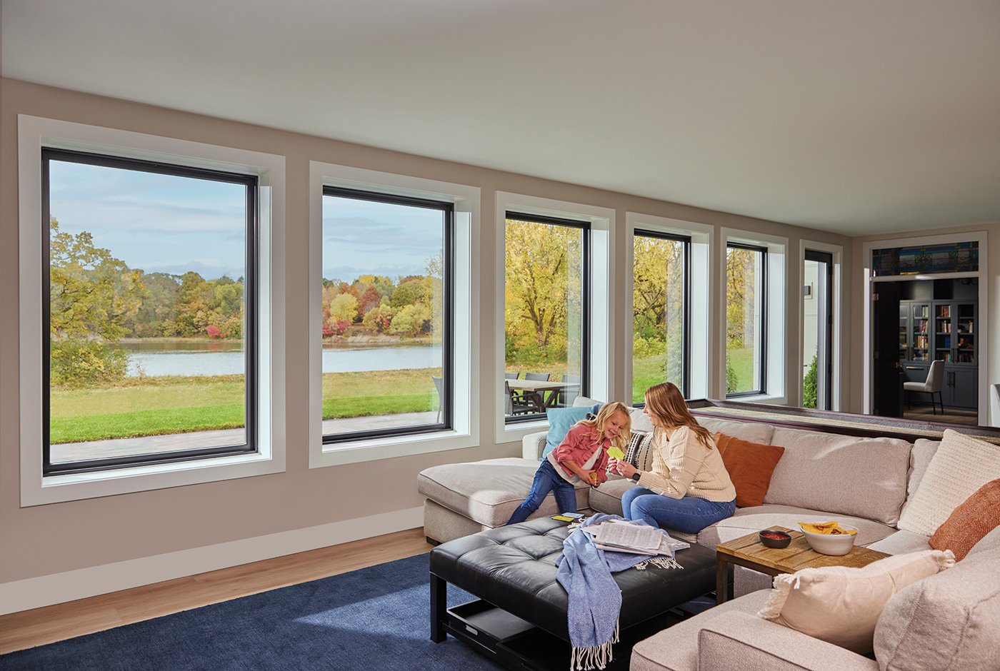 A mother sits on a sectional talking to a child in front of a row of five large picture windows that look out onto a patio with a green lawn and pond beyond.