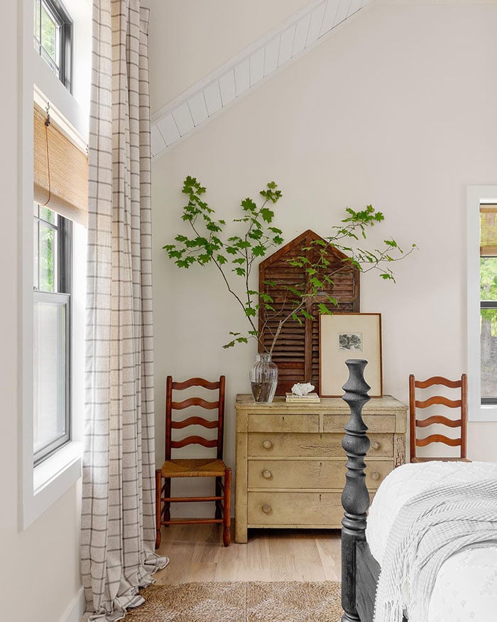 Two photos of a bedroom with warm white walls, black windows with modified colonial grilles, woven grass shades and a checked curtain, and a mix of heirloom wooden furniture.