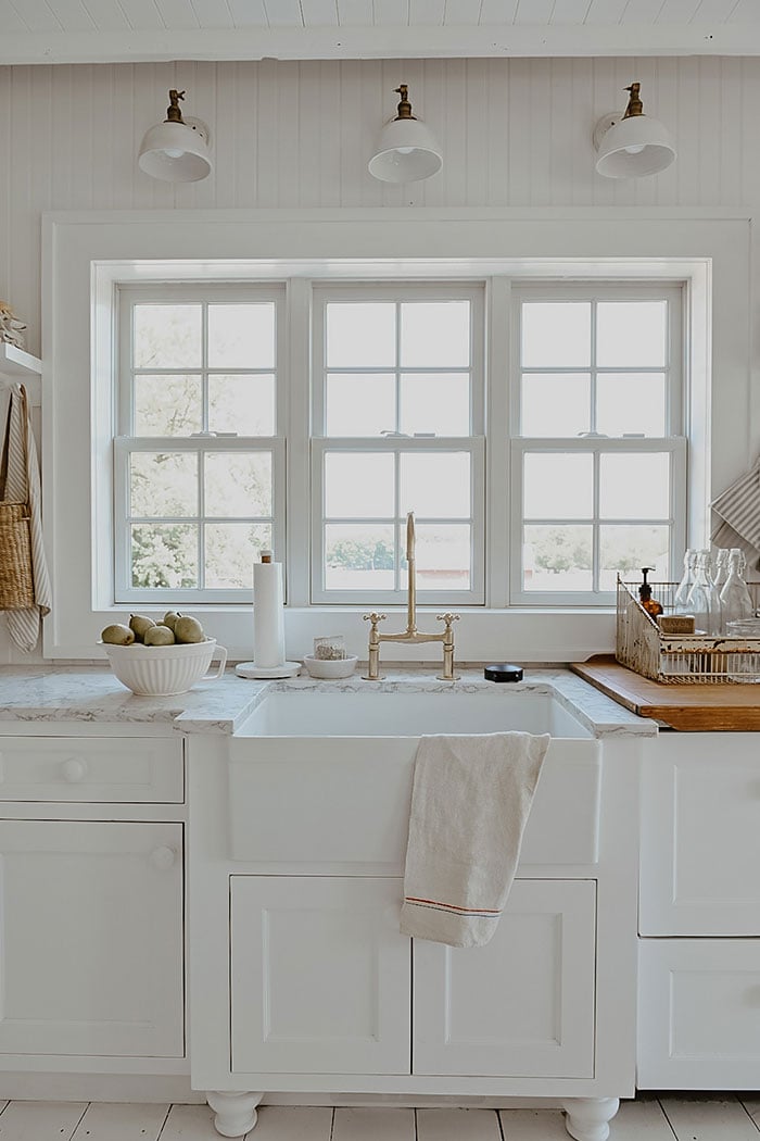 Two photos of a warm white cottage kitchen with wood-paneled walls, marble countertops, and a row of three double-hung windows above an apron sink.