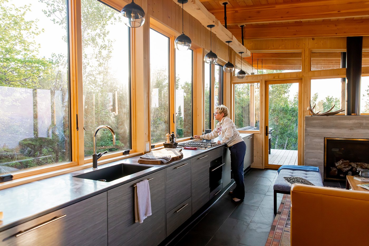 A woman stands in an open concept cabin space where a row of Andersen® 400 Series Casement Windows with contemporary profiles forms a backsplash behind the kitchen counter.