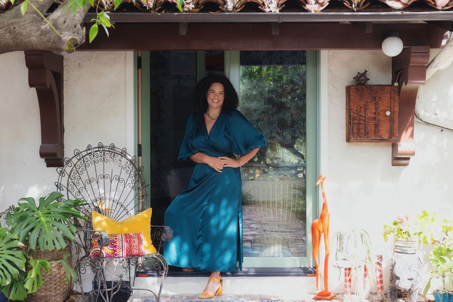 Justina Blakeney stands outside her stucco Spanish Colonial Revival home in front of an open E-Series Hinged Patio Door surrounded by potted plants, a chair, and a giraffe statue.