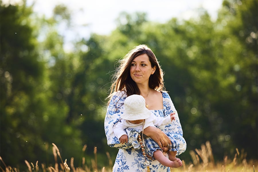 A woman wearing a white dress with blue flowers carries a baby wearing a white bucket hat as she walks across a field of golden grass with a woods behind her.