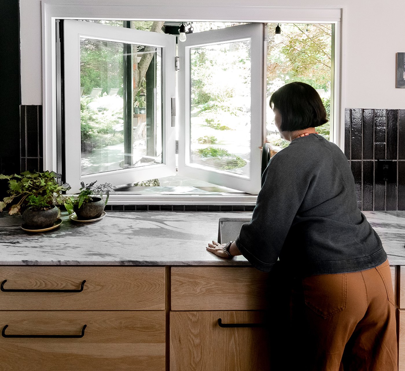 A woman opens an Andersen Folding Pass-Through Window which connects a kitchenette to a patio.