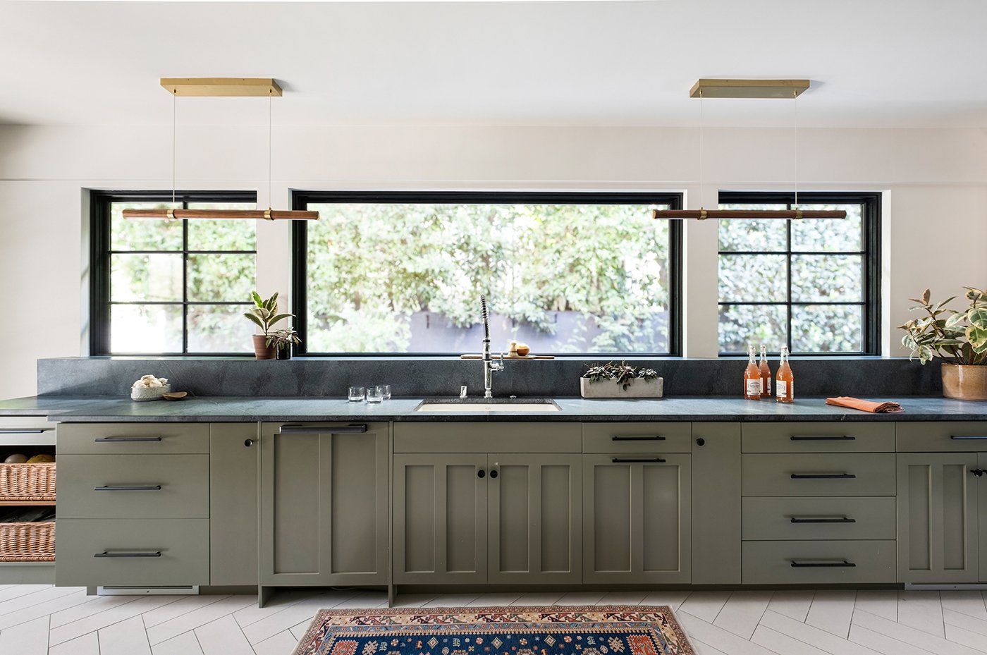 A kitchen sink and green cabinetry with black soapstone countertops above which three black-framed windows are installed.