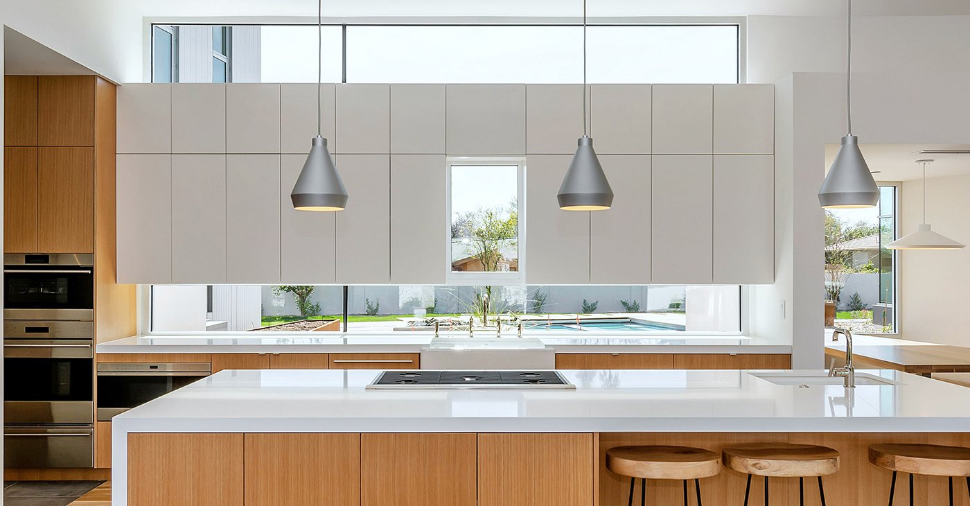 A kitchen with hardwood floors and a white countertop with stools and large windows over the sink partially covered by white cabinets. 