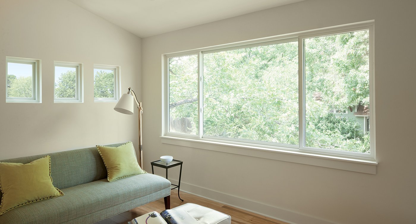 A living room with white walls and wood floors has an expansive view of the tree canopy outside through its three-sash Andersen® 100 Series Gliding Window.