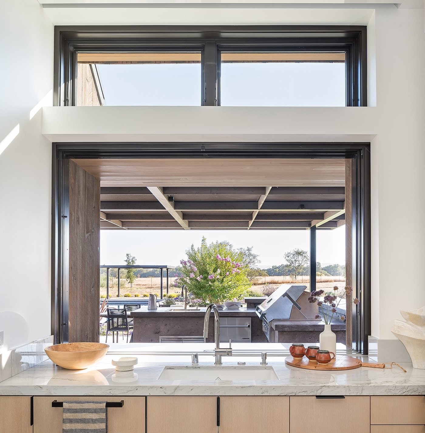 An open pass-through window connects an indoor kitchen with white oak cabinetry and marble countertops with an outdoor kitchen shaded by a large overhang. 